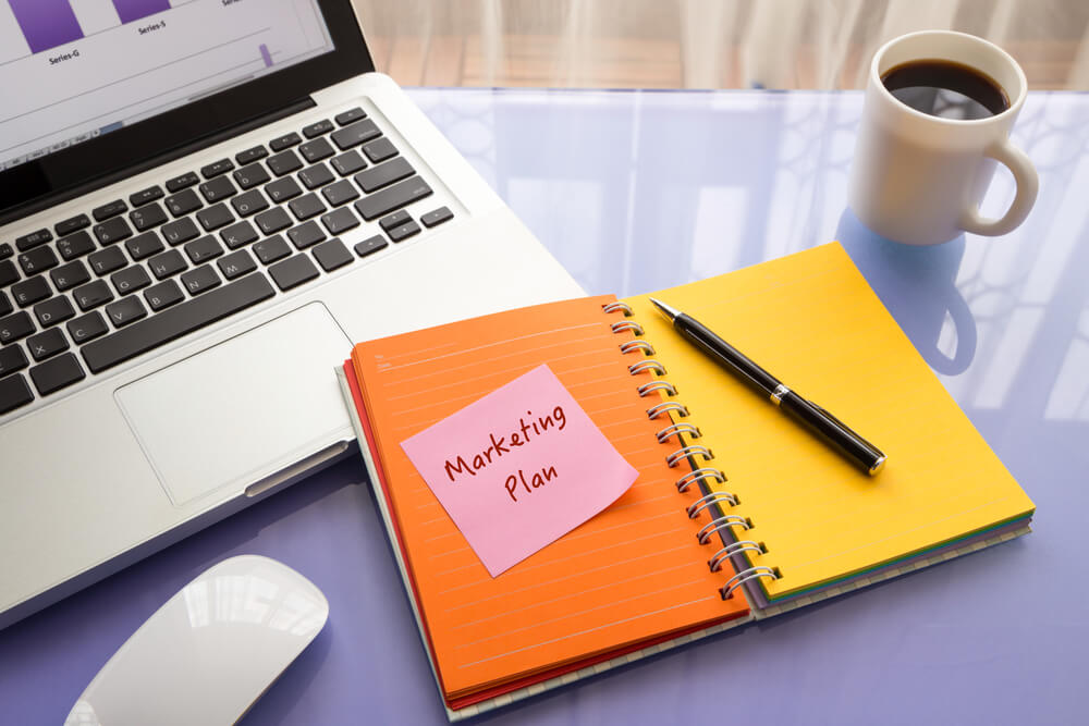 caderno em mesa junto de laptop com anotação do título plano de marketing