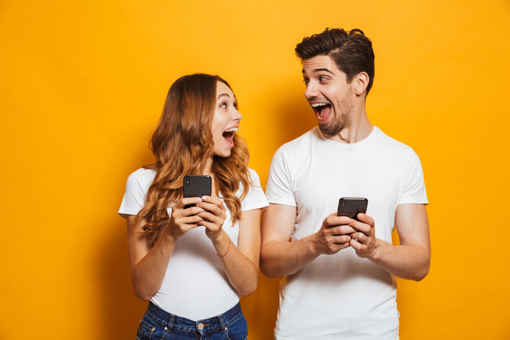casal expressivo a frente de fundo amarelo segurando smartphones