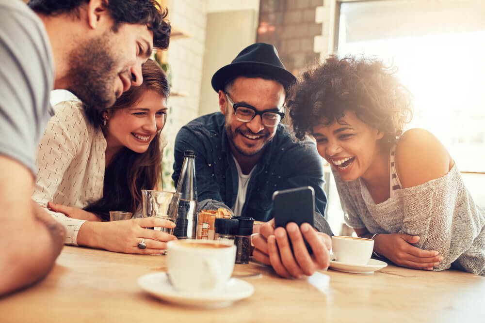 grupo de amigos sorridentes em café acompanhando algo em tela de smartphone
