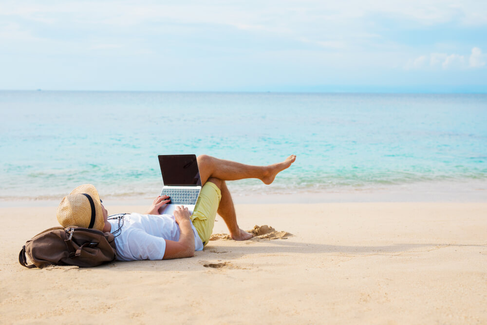 homem deitado na areia da praia com laptop no colo