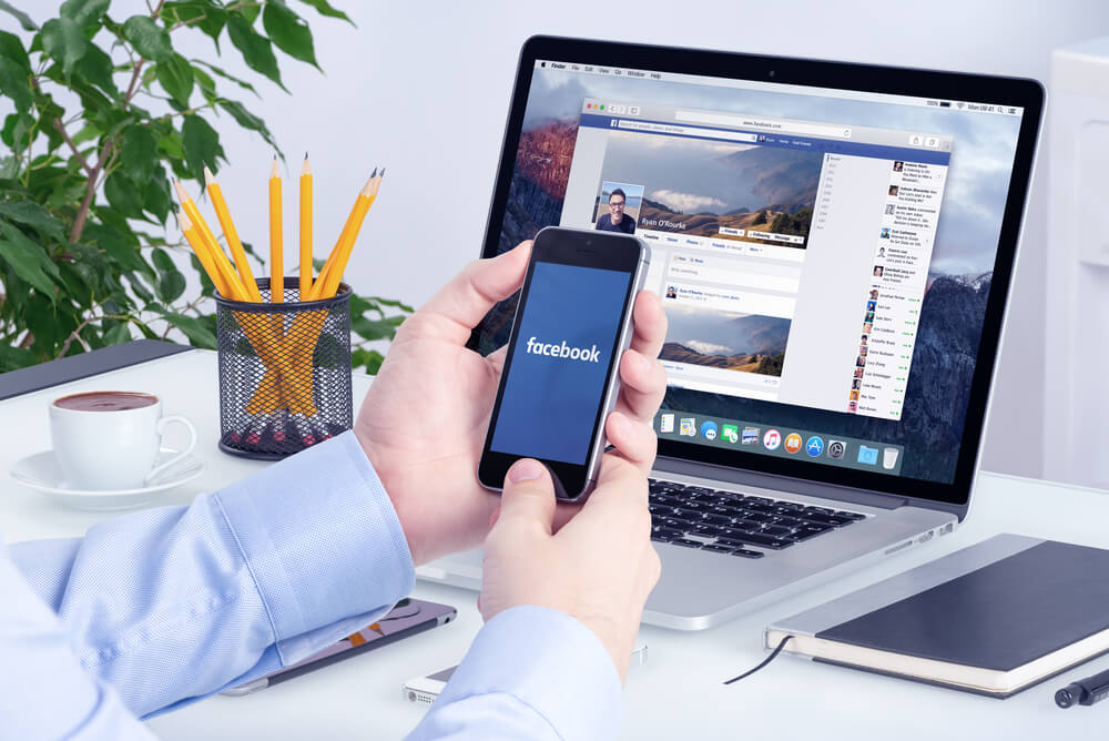 laptop em mesa de escritório com mão masculina acessando Facebook pelo smartphone