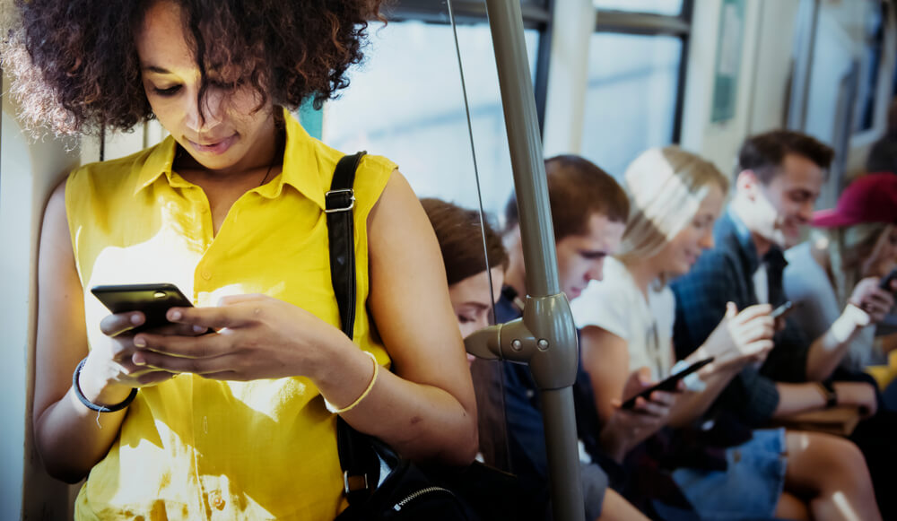 mulher em pe dentro de metro com smartphone em maos e passageiros sentados ao fundo 