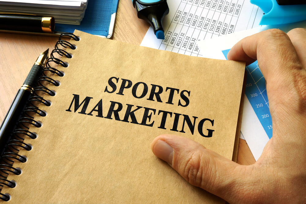 mão masculina junto de caderno sob mesa entitulado marketing de esportes
