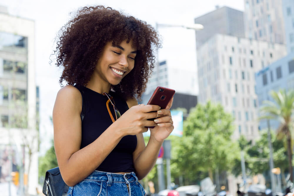 mulher sorrindo ao observar tela de smartphone em lugar publico