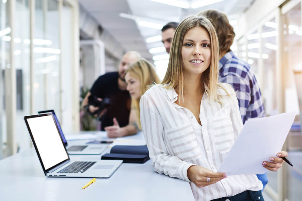 profissional na area de marketing sorrindo ao segurar papeis em frente a colegas de trabalho 