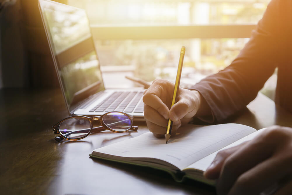 homem escrevendo redaçao publicitaria em bloco de notas com laptop e oculos de grau ao lado
