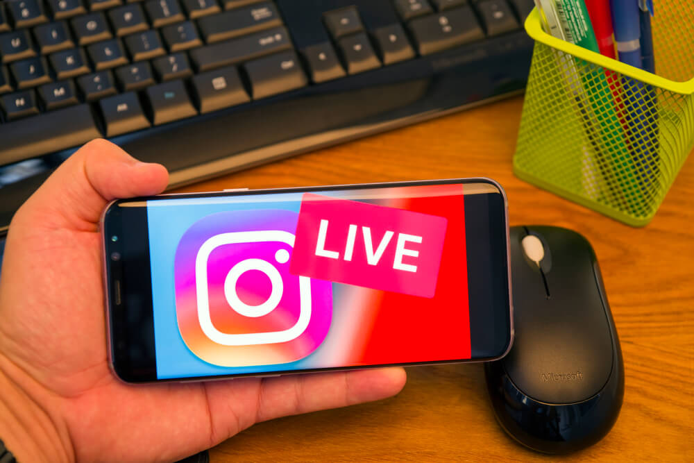 mao masculina segurando smartphone com logo do aplicativo instagram e palavra live em tela em frente a teclado de computador