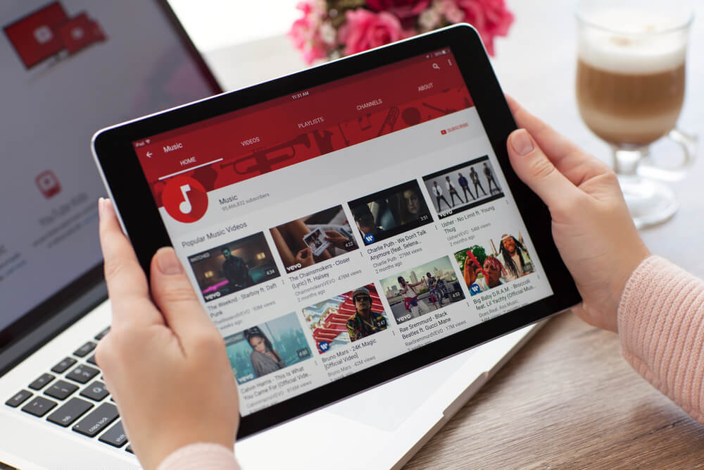 mãos femininas segurando tablet em tela do aplicativo youtube em canal sobre musicas em frente a laptop
