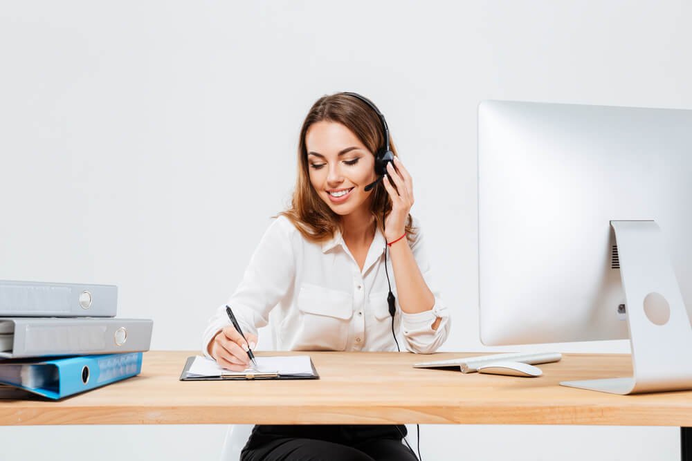 mulher efetuando vendas por telefone enquanto escreve em bloco de anotaçoes em frente a computador em mesa de escritorio
