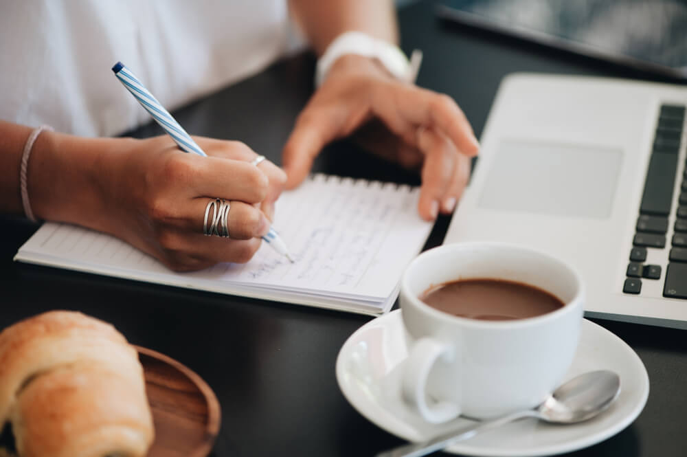 mulher escrevendo em bloco de notas com xicara de cafe e laptop ao lado