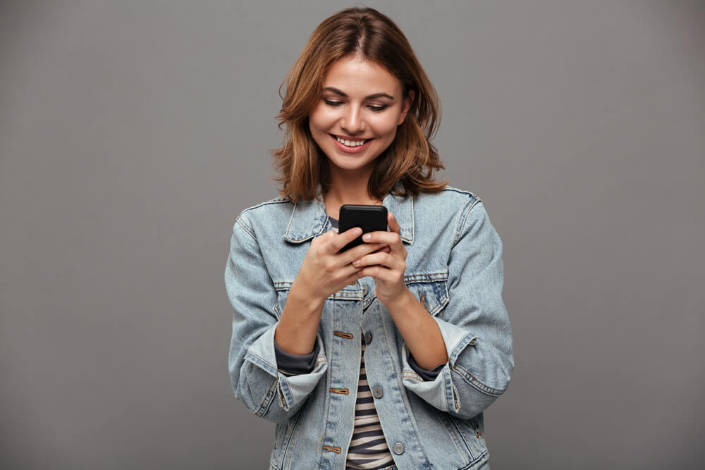 mulher segurando smartphone e sorrindo ao olhar para tela do mesmo em fundo cinza