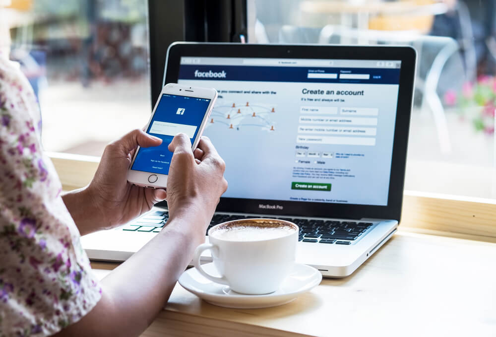 mulher segurando smartphone em frente a laptop com xicara de cafe ao lado e aplicativo facebook em tela
